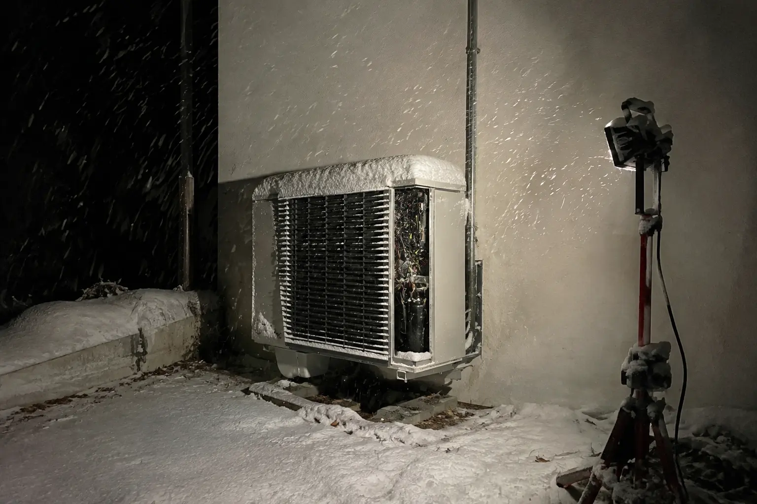Heat pump outdoor unit lit by a work light during a night call in falling snow